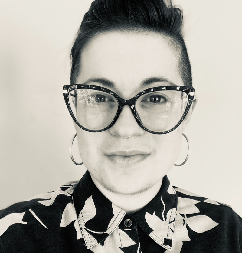 Photo of Luci Kade - black and white photo of white woman with glasses and a nose ring, wearing a black and white patterned shirt