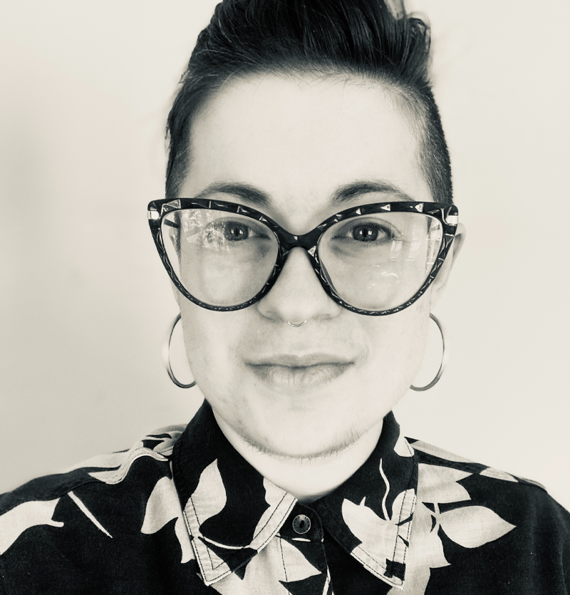 Photo of Lucas Kade - black and white photo of white person with glasses and a nose ring, wearing a black and white patterned shirt