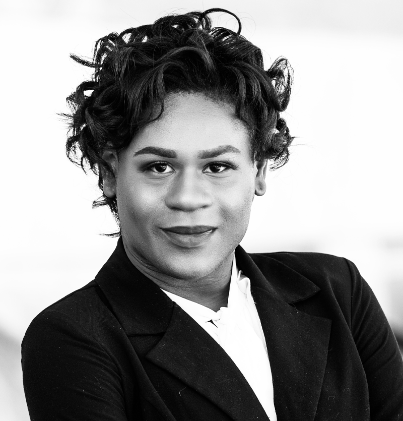 black woman wearing a white shirt and dark jacket, looking right into the camera