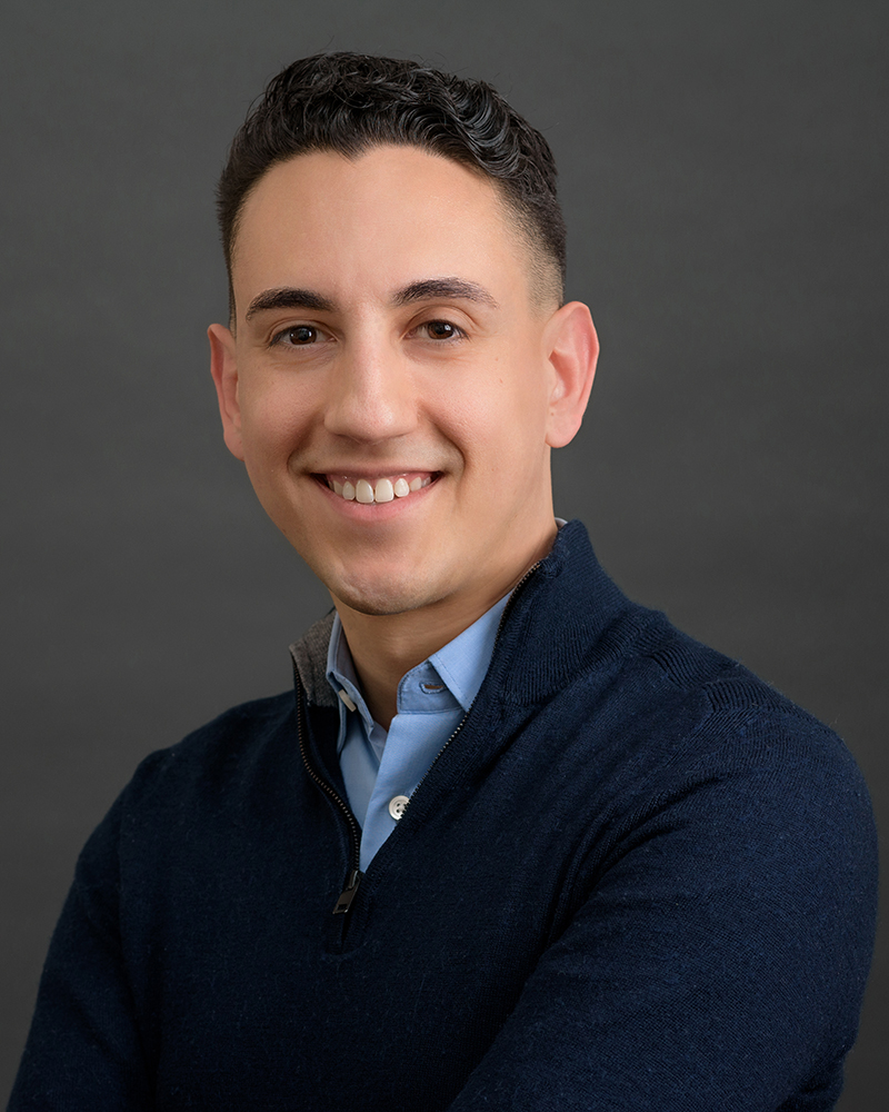 Latino man looking at the camera; wearing a light blue dress shirt under a sweater