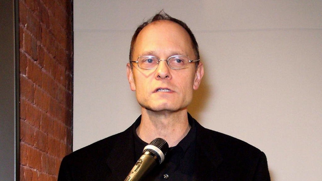 White Man With Glasses And Black Shirt In Front Of A Microphone