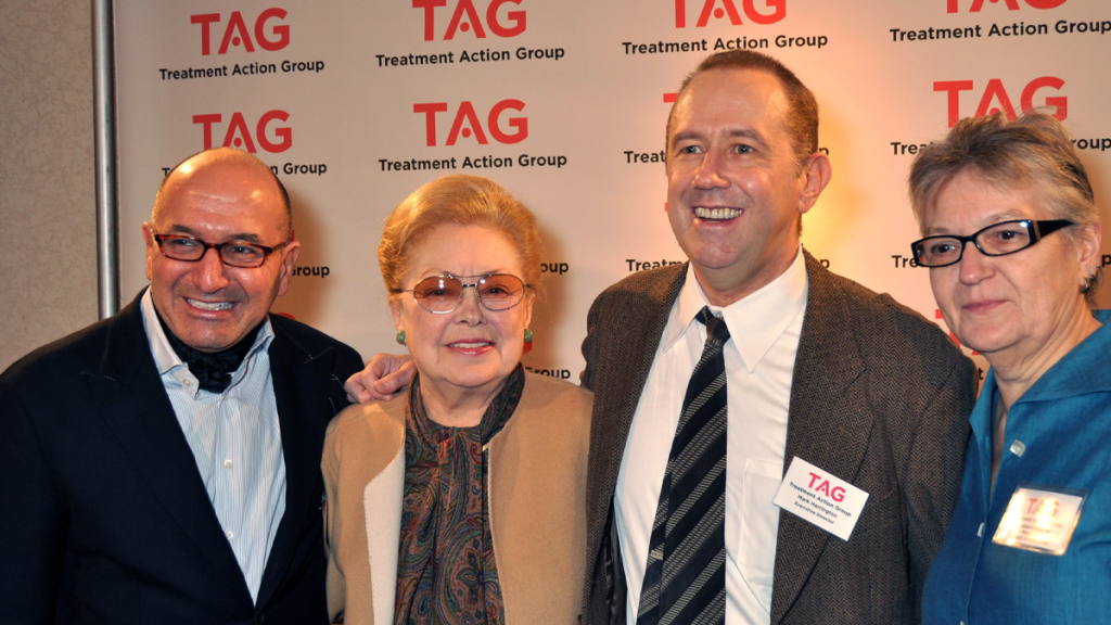 Four White People In Dressy Clothes Looking At The Camera In From Of A TAG Step And Repeat. One Has A Visible Nametage That Reads Mark Harrington