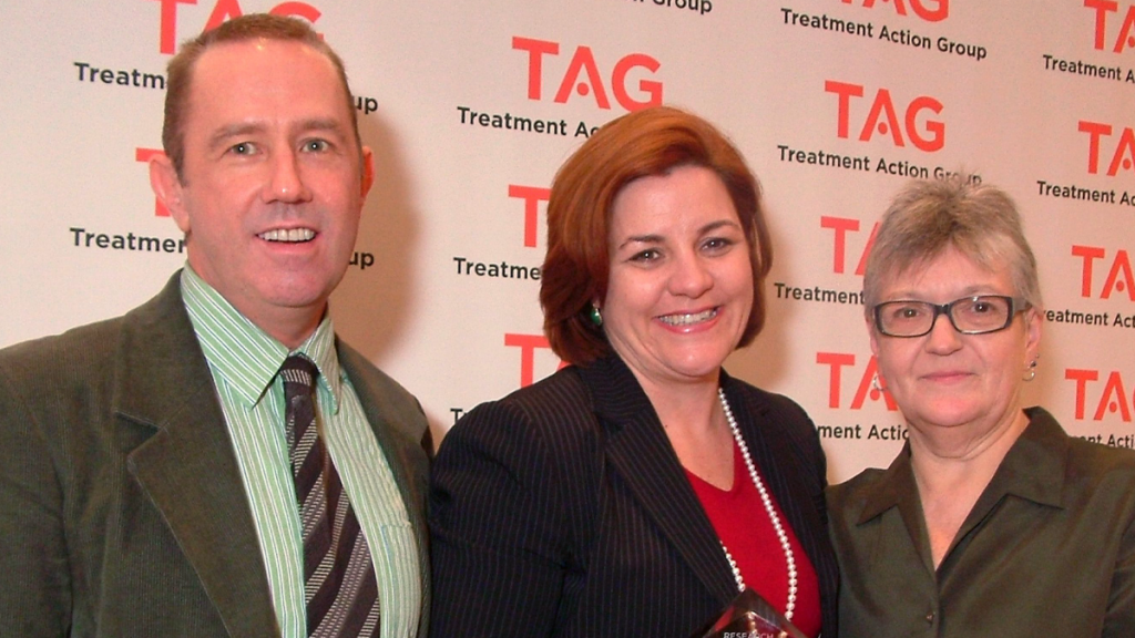 One White Man And Two White Women Smiling At The Camera, In Front Of A TAG Step And Repeat