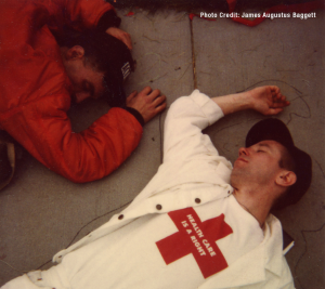 two white men lying on the ground pretending to be dead. One is wearing a t-shirt that clearly reads "Health Care Is a Right"