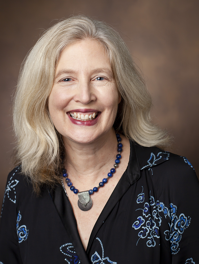 Photo of smiling blond woman; wearing a black dress with a blue print and a black and blue necklace