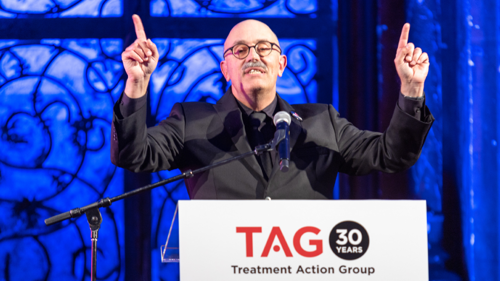 Man Wearing Glasses On Stage, Raising Each Index Finger; At Podium With TAG 30 Years Podium Sign