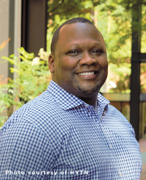 Black man smiling at camera, wearing a blue and white gingham shirt