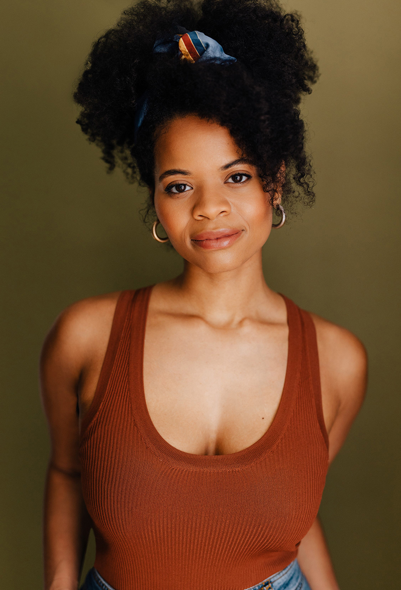 youngish black woman smiling, has with her natural hair up, is wearing a copper tank top and jeans