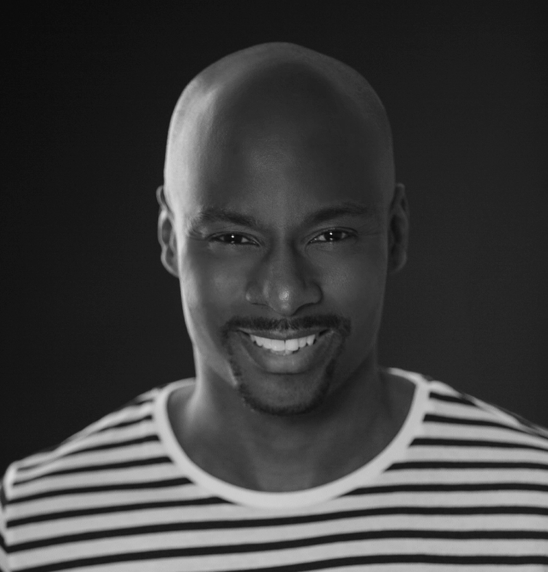Black and white photo of a black man with a goatee, smiling, and wearing a striped collarless shirt against a dark background