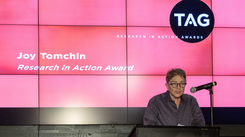 Older Woman At Microphone Giving A Speech. The Screen Behind Her Says "Joy Tomchin, Research In Action Award"
