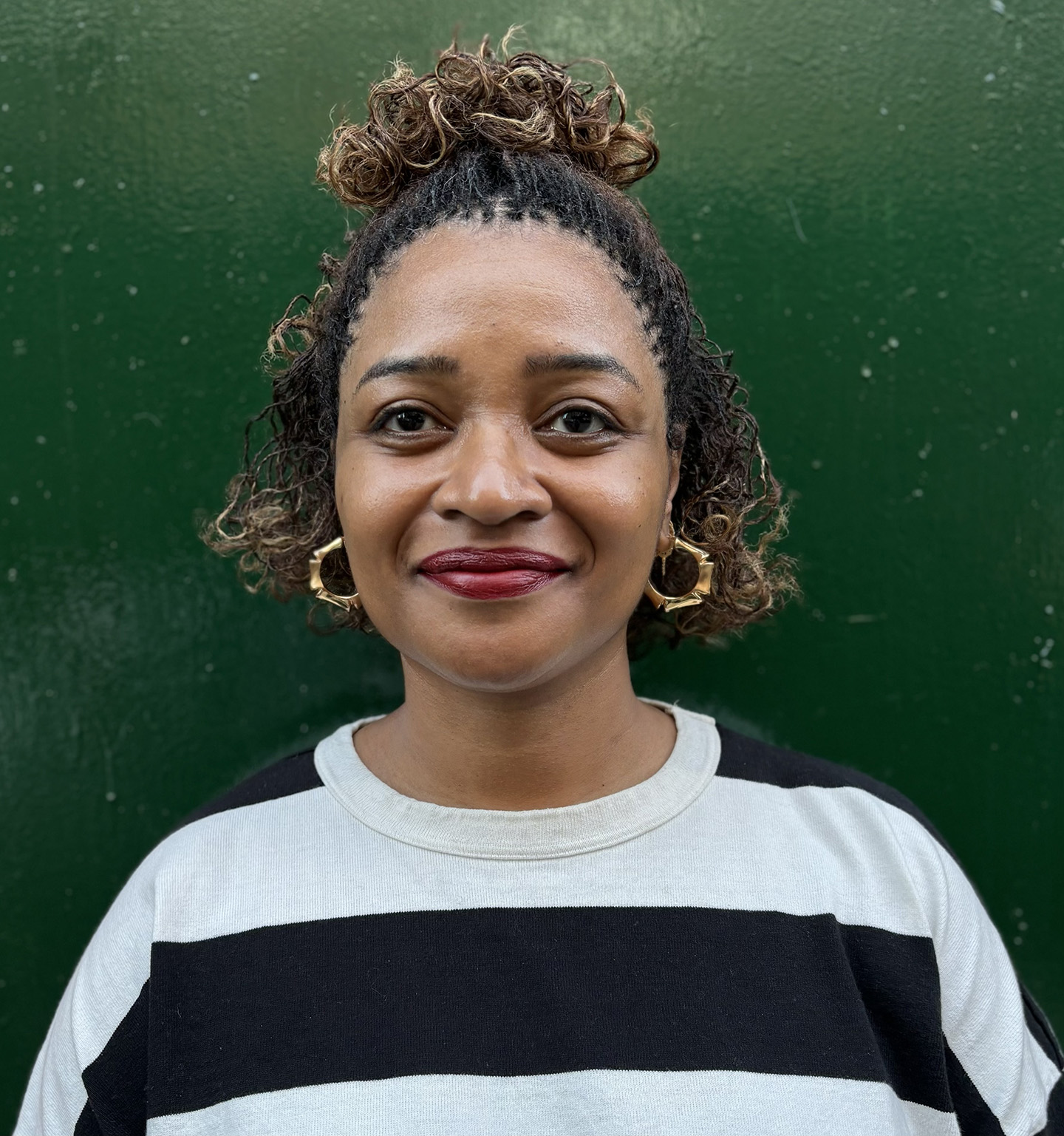 woman of color wearing a black and white striped top, gold earrings, and smiling, against a green background